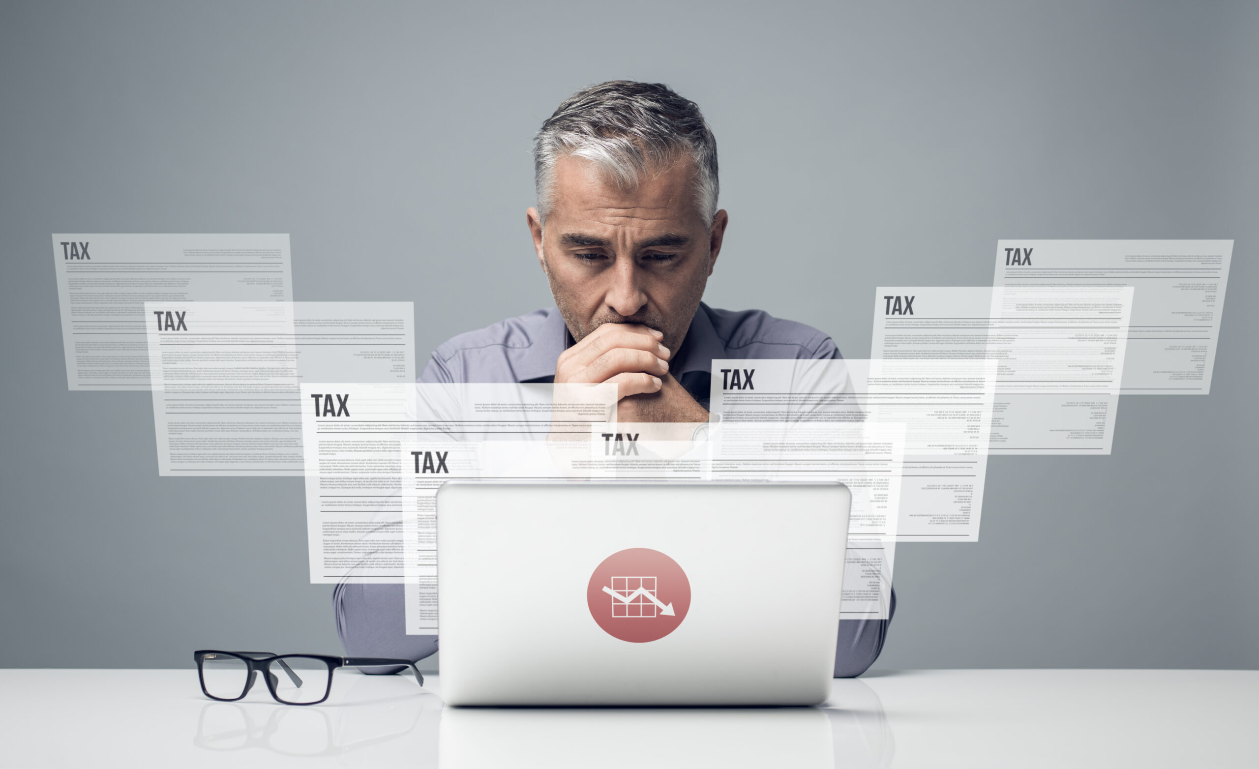 A man appears stressed while reviewing multiple tax documents on his laptop, emphasizing the complexities of tax preparation.
