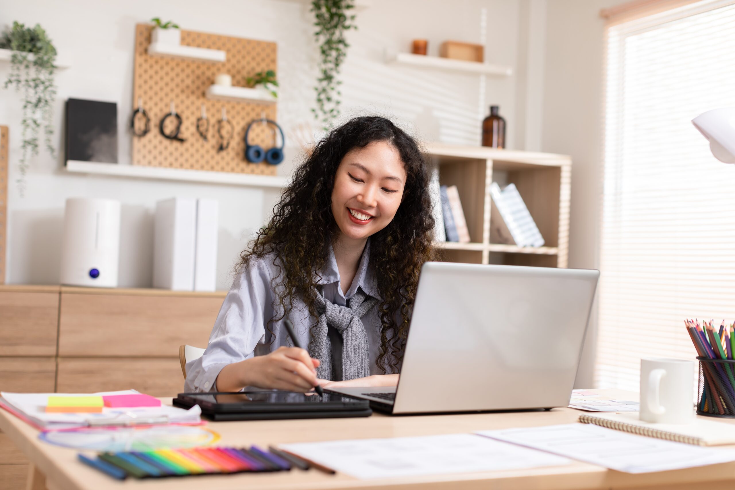 A smiling woman works on a laptop and graphic tablet in a modern, organized workspace, filled with art supplies and plants, emphasizing creativity and productivity.