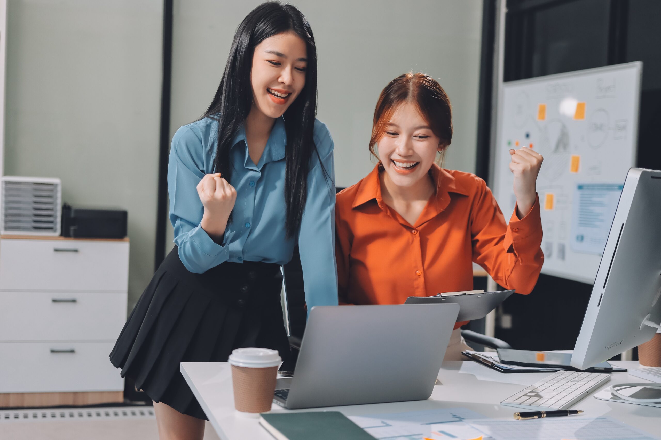 Two young women celebrate success in a modern office, smiling and gesturing excitedly over a laptop, surrounded by work materials.